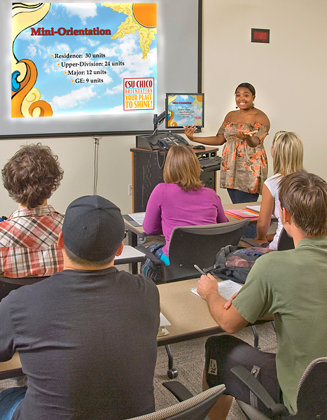 Woman standing next to a smart podium, leads students in a mini-orientation.