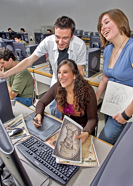 Three students laugh while looking at a computer. The two women hold different drawings of the same character. 