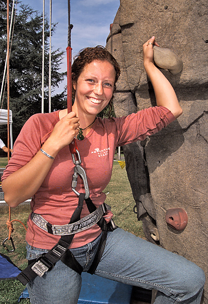 Woman wearing climbing harness smiles with left hand and leg up on rock wall grips.