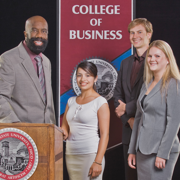 Four people smile, stand in front of College of Business banner against black background.
