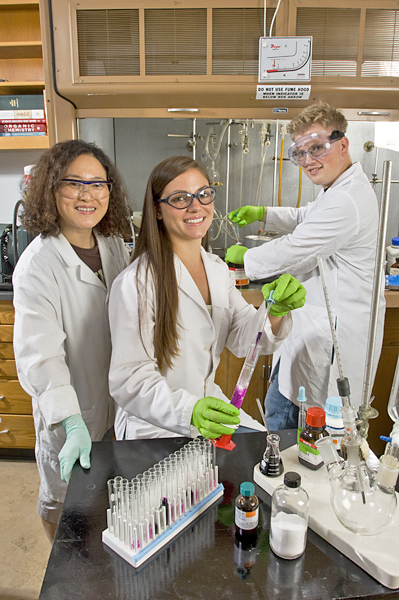 Three students wearing lab coats and gloves smile at the camera. Two hold laboratory materials—graduated cylinders and eye-droppers.