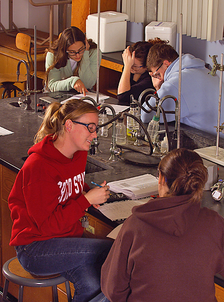 Five students in two groups sit at a lab table with open books and notes. 