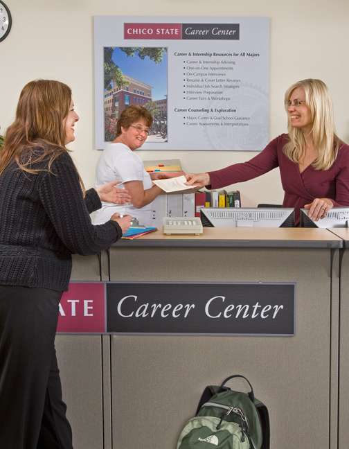 Female career center staffer standing behind a counter hands a paper to woman on this side of the counter. Both women smile and so does a woman in the back who flips through a binder.