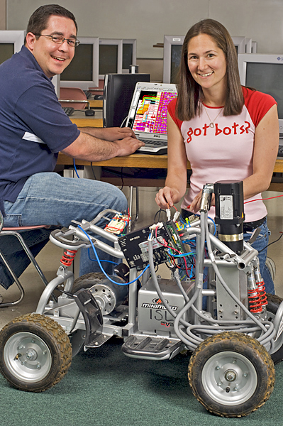Man sits at computer, smiles at camera, while woman works on a robotic vehicle.