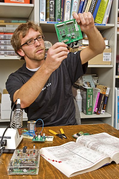 Man wearing glasses sits at table holding up a circuit board with reference book on desk in front of him.