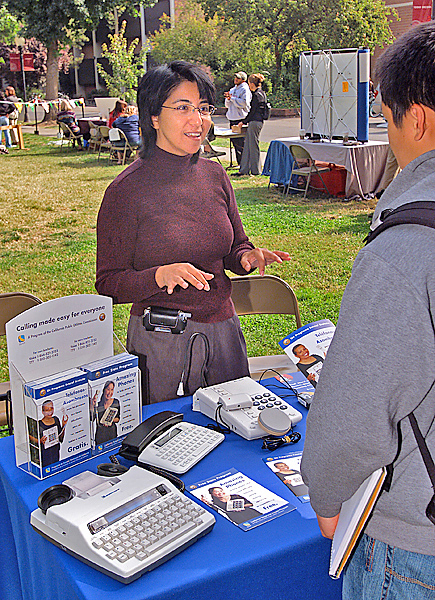 Woman (center) speaks across table to student about sevearal TDD receiver models displayed.