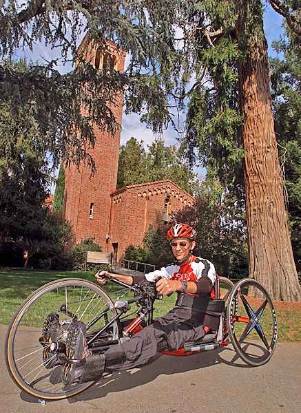 Man, facing left, wearing helmet and cycling gear, sits in hand-propelled three-wheeled bike.