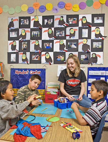 Woman sits on table where three young boys play with clay and draw with colored pencils.