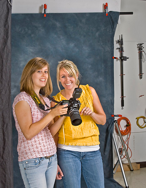 Two women stand in a photography studio looking at a camera that the woman on the left is holding.