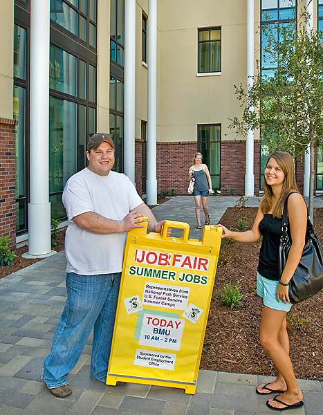 A man and a woman set up a yellow sandwich board with the words "Job Fair" in red capital letters across the top.