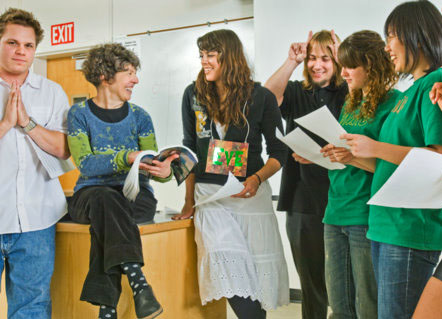 Five students, each holding a sheet of paper, surround older woman who holds a book. One student makes horns next to his head, while another clasps his hands together in a praying position. 