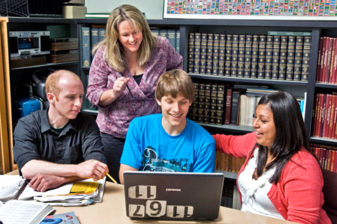Three people sit at a table around a laptop, while one woman stands over the group peering at the computer screen. They are in some sort of library.
