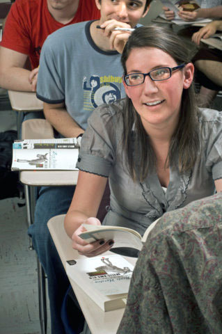 Woman with glasses sits at desk in a classroom holding a book open and smiling as she looks to the left.