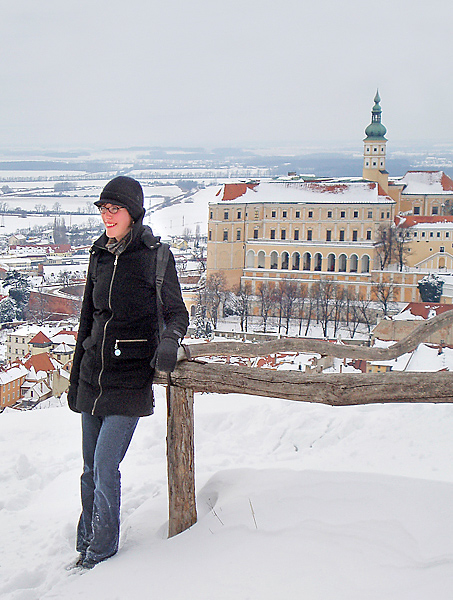 Woman (left) facing left, stands smiling on top of snow covered hill with city in background.