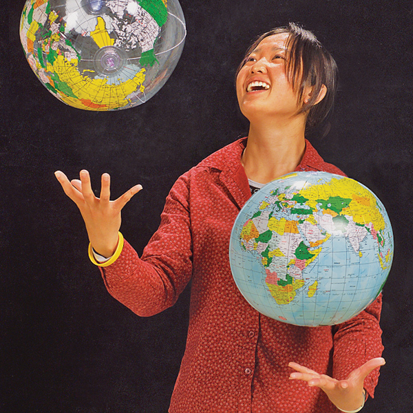 Woman in red jacket, wearing yellow wrist band juggles two inflatable globes in front of a black background.