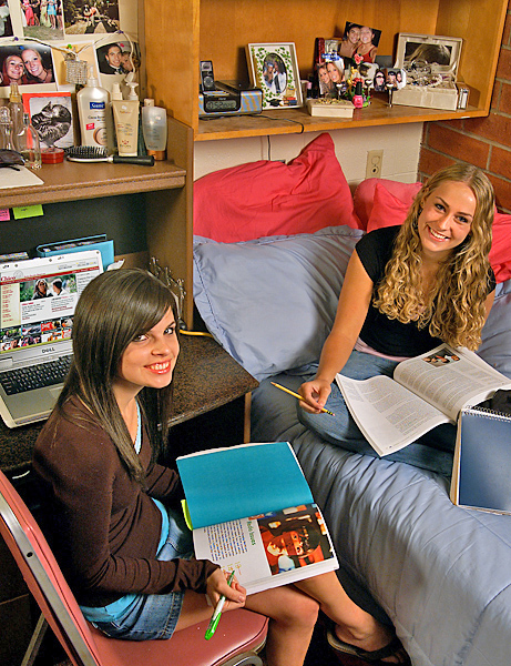 Woman (left) sits in chair facing woman sitting in bed; both smile and hold books.