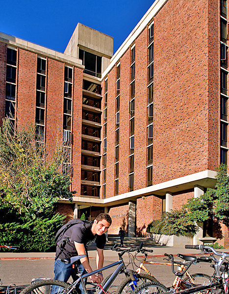 Man with backpack leans down to lock up bike; Whitney residence hall towers above.