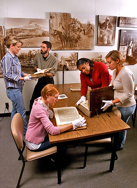 Five people all wearing white gloves are in an archival setting, delicately perusing historical documents. One woman sits at a table where two other women flip through a huge book.