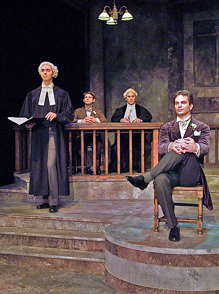 Four men hold court in a theatrical period performance. Two men wear white wigs and black robes, one standing and holding a book. One man sits "on the witness stand" alone.