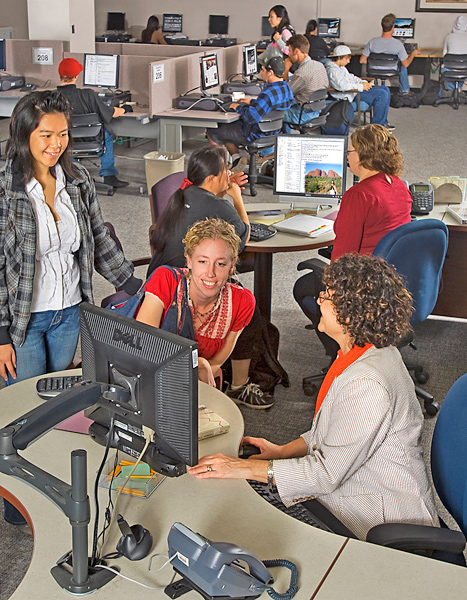 In a computer lab, one woman sits behind a desk and helps two women at her desk. One stands while the other woman sits and leans over to touch the screen.