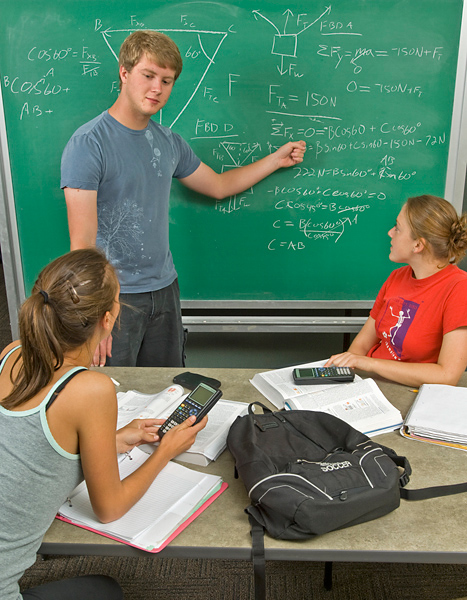 Man stands at chalk board, points to math equations; two women with calculators and books sit on opposite sides of a table listening.