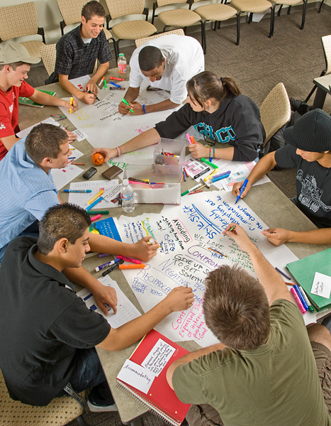Seven men, one woman seated around a table create colorful posters using markers.