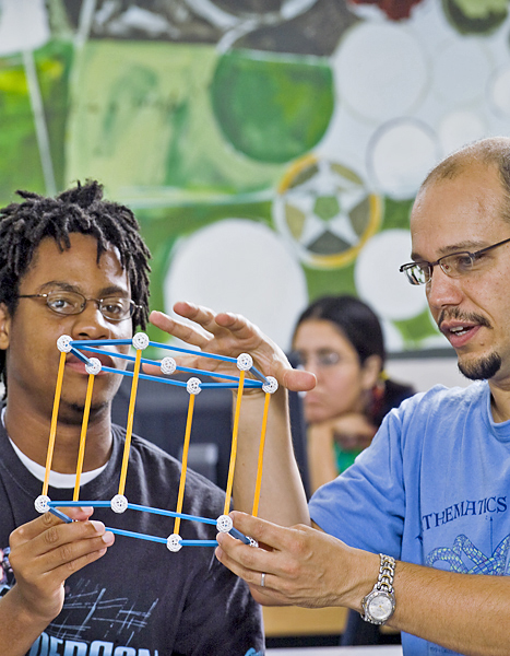 Two men, each wearing glasses, hold a 3D model as man on right gestures to the model during their discussion.