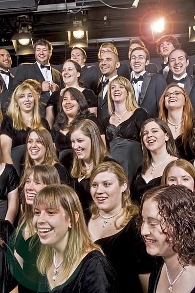 A large group of music students, all dressed in black formal wear, share a laugh on stage. Stage lights illuminate the group.