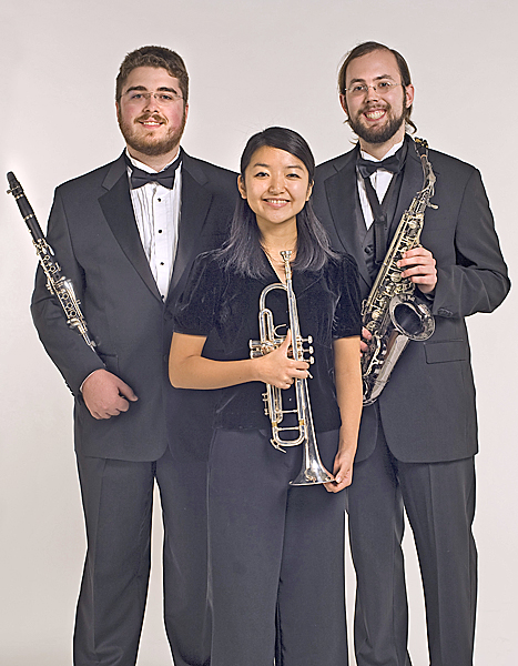 Three music students, all dressed in black formal wear, stand in front of a grey background and hold their instruments; a flute, trumpet and saxophone, respectively.