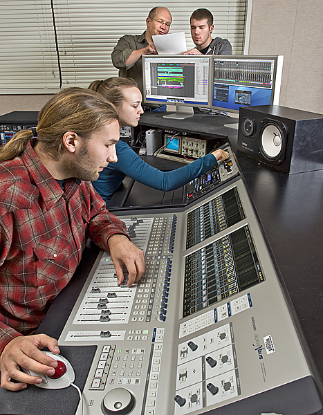 Man and woman, both with pony-tailed hair, make adjustments to a huge sound board. In the background, two men study a sheet of paper using a computer monitor for support.