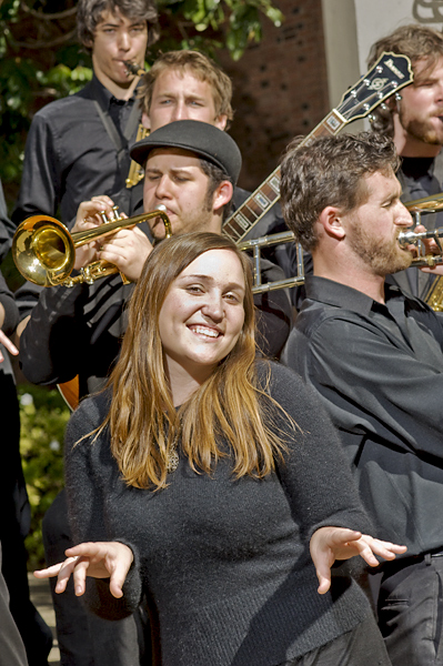 A smiling woman wearing a black sweater, takes center stage in this photo as a band with trumpet, saxophone, guitar, and trombone play behind her.