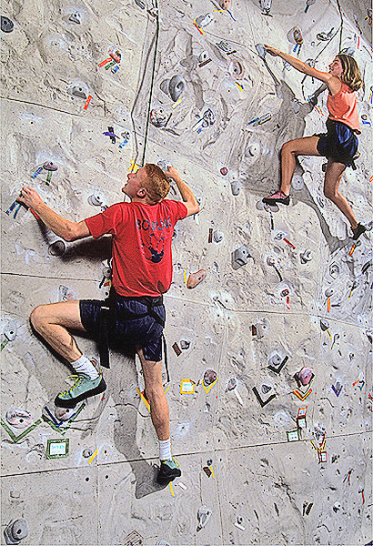 Man wearing red shirt and black shorts climbs a rock climbing wall looking upward on the left as a woman in an orange shirt and blue shorts climbs just a bit higher on the right.