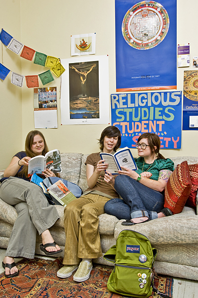 Three women sit on a couch. Two share a book, one reads alone. Religious artifacts decorate the wall behind them.