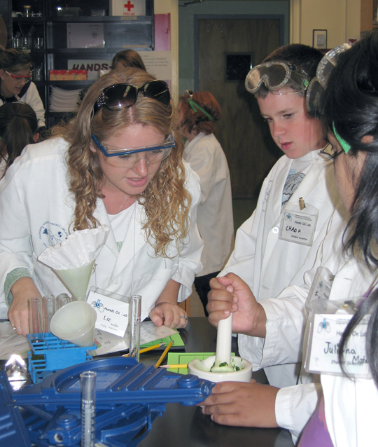 Science Ed student (left) leans in to inspect one of three elementary students' work on table.