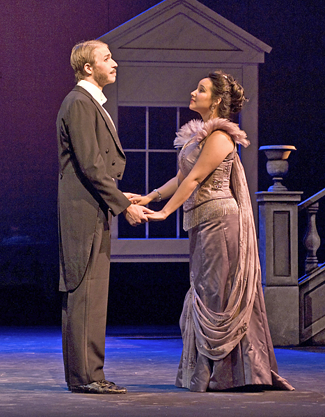 Man and woman, dressed in early 20th-century formal wear, hold hands. Man stares off, woman stares at him.