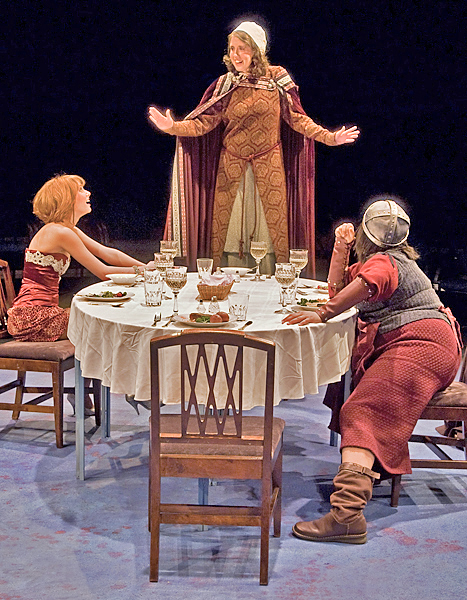 Three women in mid-eval garb are around a table set with glass-, silver-, and plateware. One woman stands on a chair at head of table. One woman in a dress smiles and gazes up at standing woman and another woman leans with one leg out and elbows on the table staring up at the standing woman.