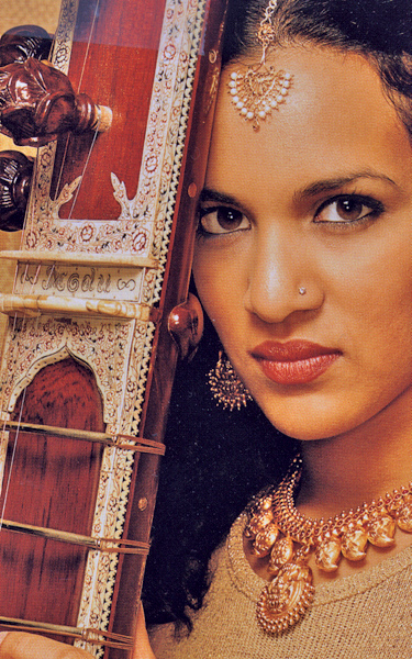 Female global music practitioner, with matching multifaceted necklace, earrings, and head jewelery, poses next to an exotic instrument that touches her forehead.