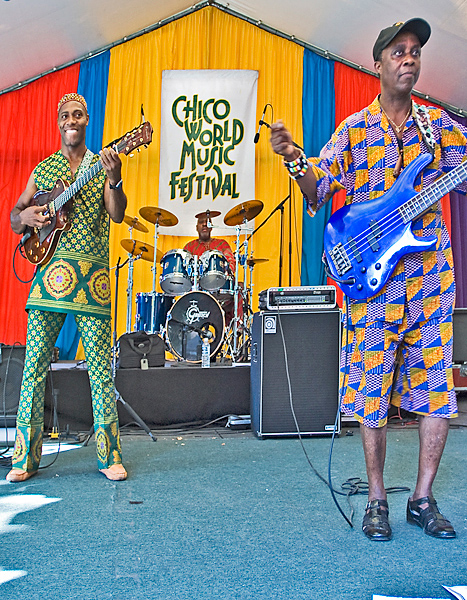Band members, dressed in brightly colored ornate-patterned clothing warm up before a music set. Two guitarists, with instruments slung over-shoulder, look out into crowd while drummer behind them looks on as well.