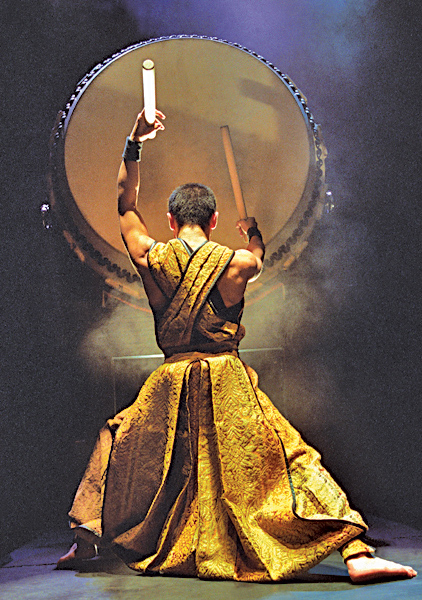 Man in traditional clothing, stands with his back to camera, against a smoky black background, ceremoniously hammers on a huge drum.