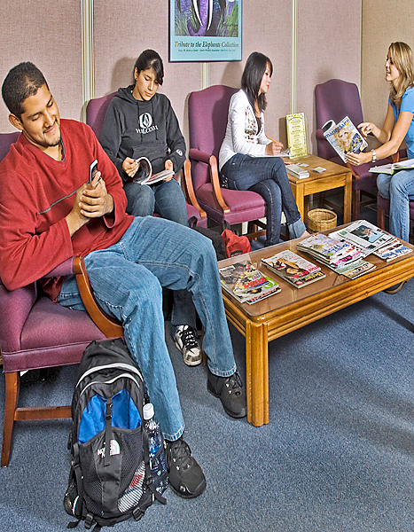 Several students sitting in lobby; one man looks at his cell phone, one woman reads magazine, two other women chat while filling out forms.