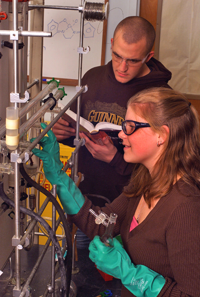 Man holding book watches as woman makes an adjustment to a machine wearing gloves and goggles.