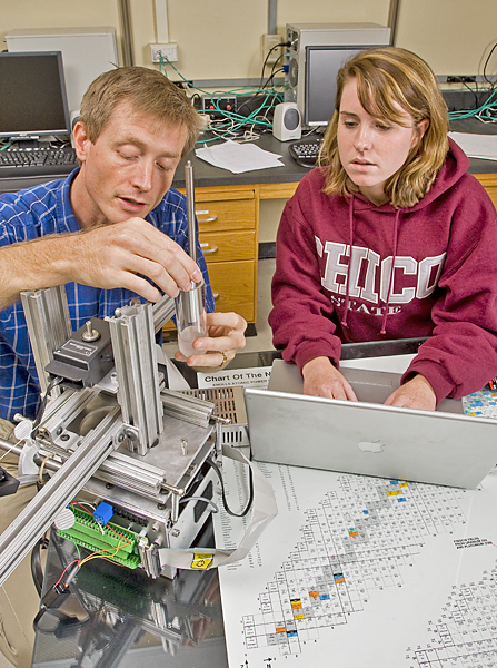 Man works on some device being used for an experiment. Woman sits next to him observing and typing on a laptop computer. 