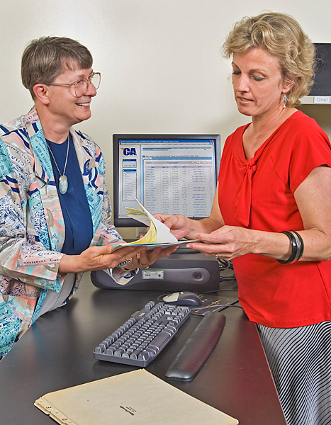 Two women, standing on opposite sides of a counter, hold a stack of papers together. One on right flips through the stack while the woman on the left smiles.