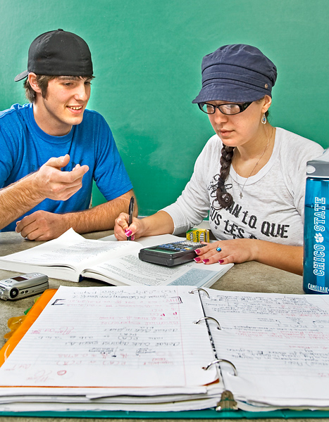 Woman wearing a hat and glasses holds a pen to paper while looking at an open book, open binder and calculator. Man in backwards cap sits next to her and gestures with his hand out.