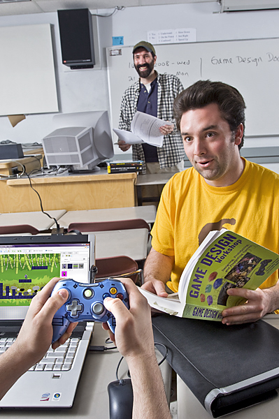 Man in yellow shirt holds book on game design while bearded man with hat smiles in background.