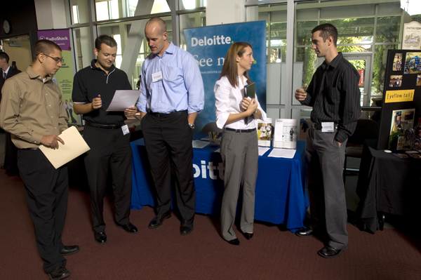 A group of five people stand in front of a table and standing banner with "Deloitte" in white letters against a blue background. On the left, three men look over a document that a man in black holds. The other two men, on the in a brown shirt and glasses, on the right a man in a blue shirt flank the man holding the paper. On the right, a woman and man look as though they are engaged in conversation.