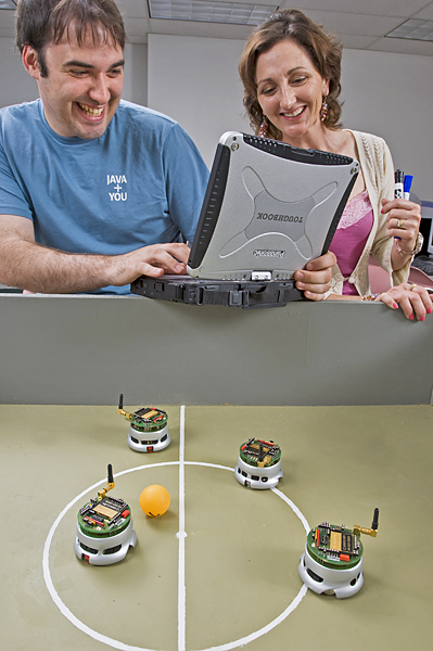 Man with laptop computer stands next to woman holding markers while both watch a four small robots "play" soccer with a ping pong ball.