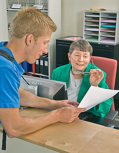 Male student leans on counter pointing to papers that he holds out to seated woman on the other side of the counter who puts on her glasses as she begins to look at the papers.