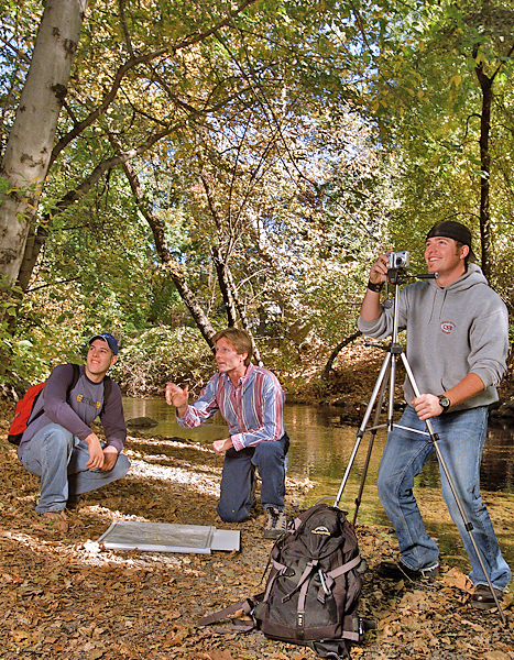 Two men kneel near creek looking at something to the left. A third man steadies a tripod with one and ready to snap the photo of something out of frame to the left.