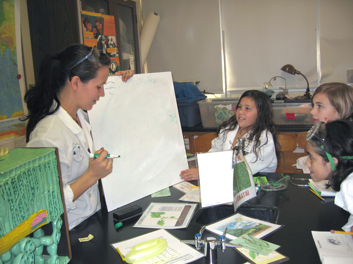 Woman (left) holds white board poised to write as three young girls look on, ready to answer.